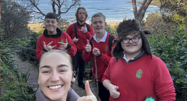 a teacher with 4 students at the beach all smiling and giving a thumbs up