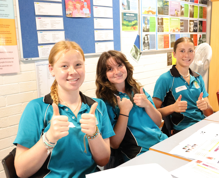 3 students giving a thumbs up sitting at a desk in a classroom
